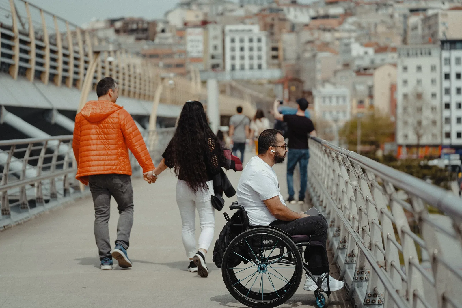 A couple and a man in a wheelchair enjoying a walk on a city bridge with urban backdrop. First accessible trips run late for one arithmetic reason