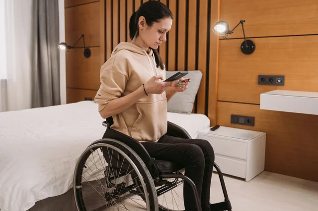 Woman in wheelchair using smartphone in a modern wooden-paneled bedroom, creating a calm atmosphere. 7 things to weigh up on accessible weekend destinations