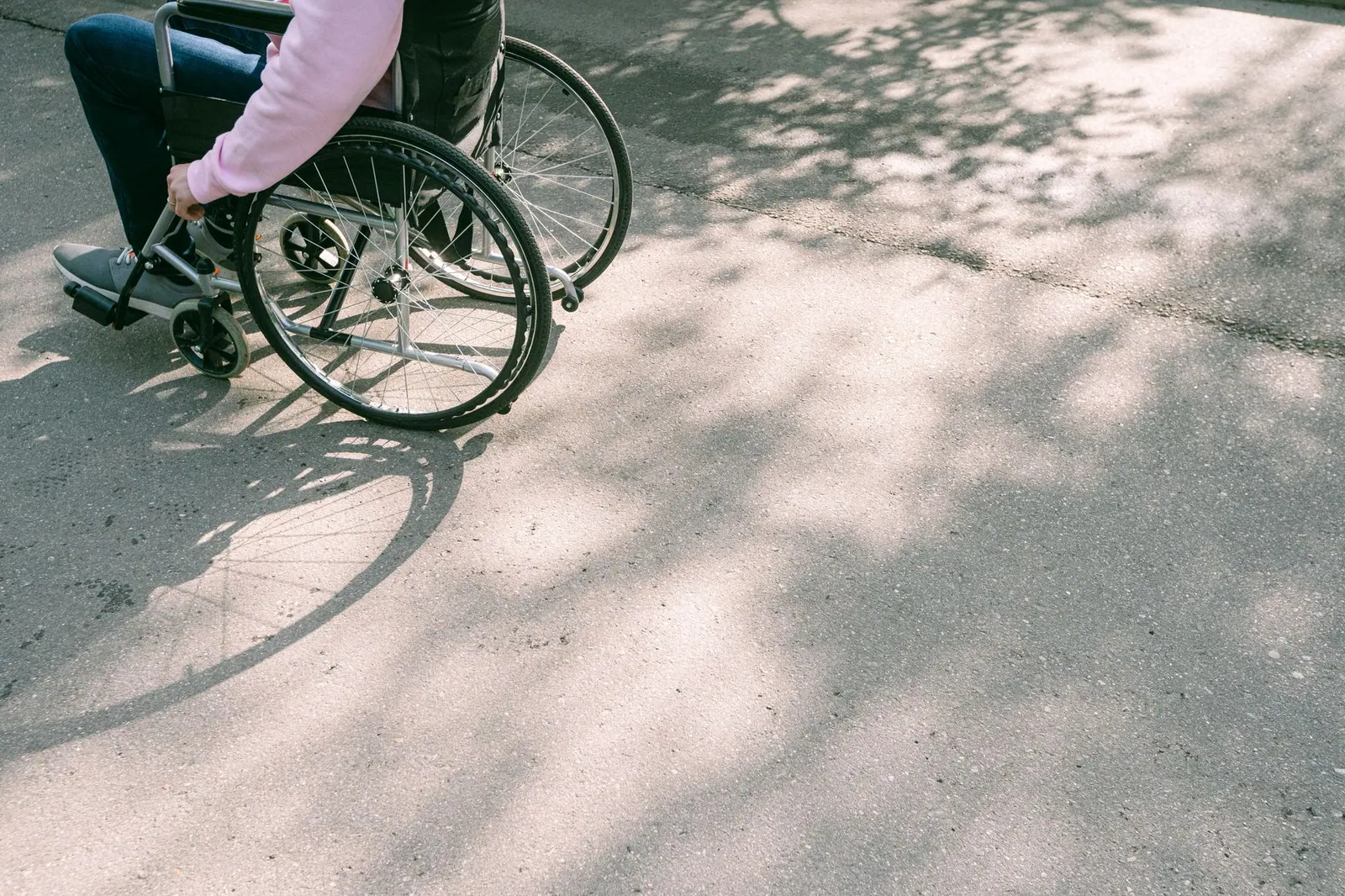 A person in a wheelchair navigating a sunlit road with tree shadows. The image captures a moment of mobility and independence.