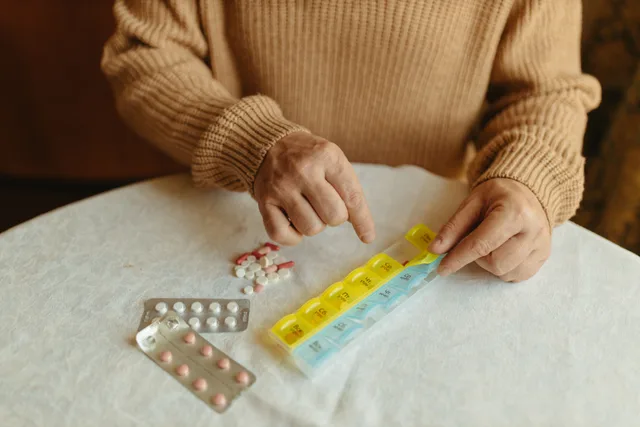 Senior adult organizing daily medications using a weekly pill organizer on a table indoors. Packing for an accessible weekend: redundancy beats extra volume