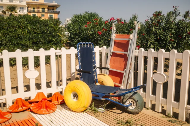 Beach wheelchair next to lifeguard equipment by a white fence on a sunny day. How to time an accessible weekend trip around crowds and heat