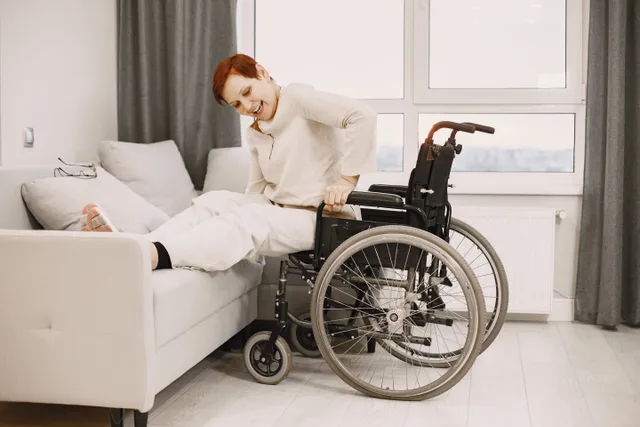 A woman with dyed short hair smiles while transitioning from a wheelchair to a couch. A budgeting checklist for accessible travel that skips fuzzy averages