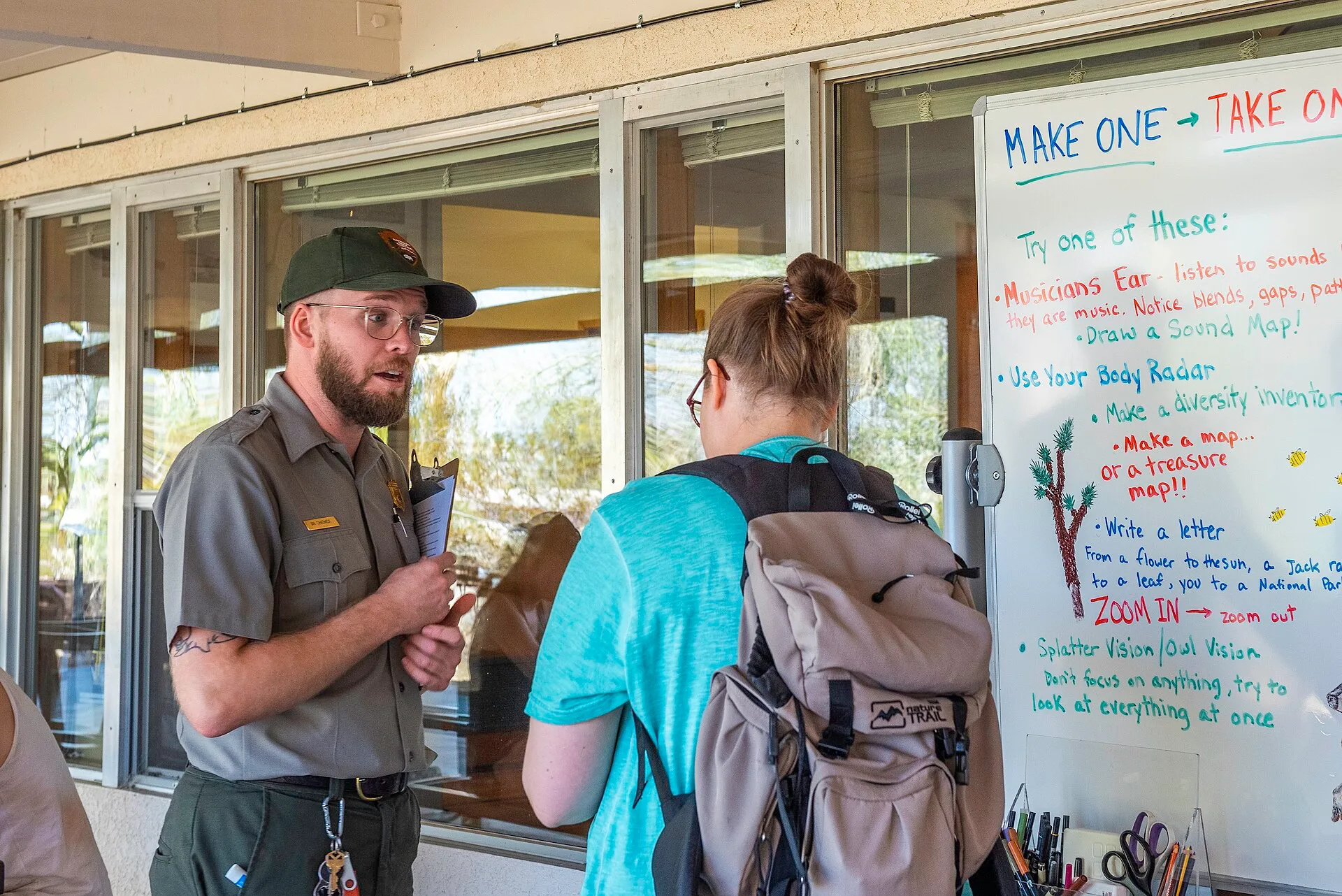 NPS / Emily Hassell Alt text: A park ranger talks with a visitor outside of the Oasis of Mara Visitor Center in front of a table of drawing supplies. Where accessible weekend guides come from: rangers, not travel agents