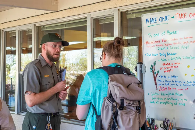 NPS / Emily Hassell Alt text: A park ranger talks with a visitor outside of the Oasis of Mara Visitor Center in front of a table of drawing supplies. Where accessible weekend guides come from: rangers, not travel agents