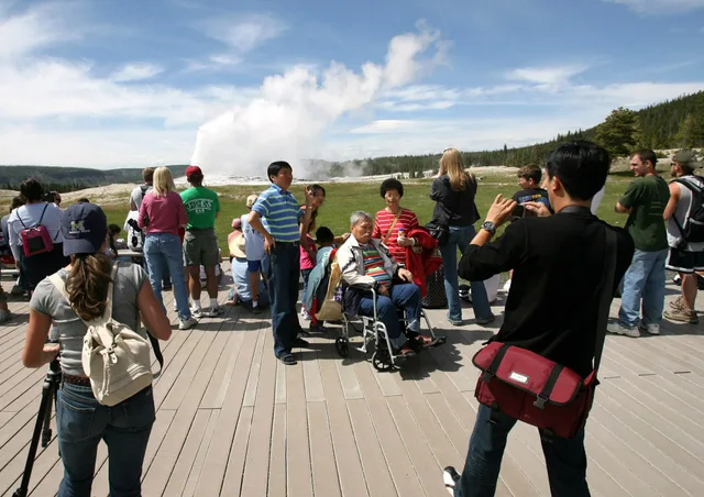 People on the boardwalk are posing for pictures in front of Old Faithful Geyser as it erupts. Visitors with wheelchair photographing an Old Faithful geyser eruption; Jim Peaco; June 25, 2008; Catalog #18744d; Original #RD7Y0865 Keywords: 18744d; thermal features in yp; yellowstone national park; exp. Six wheelchair accessible trip shapes that work reliably